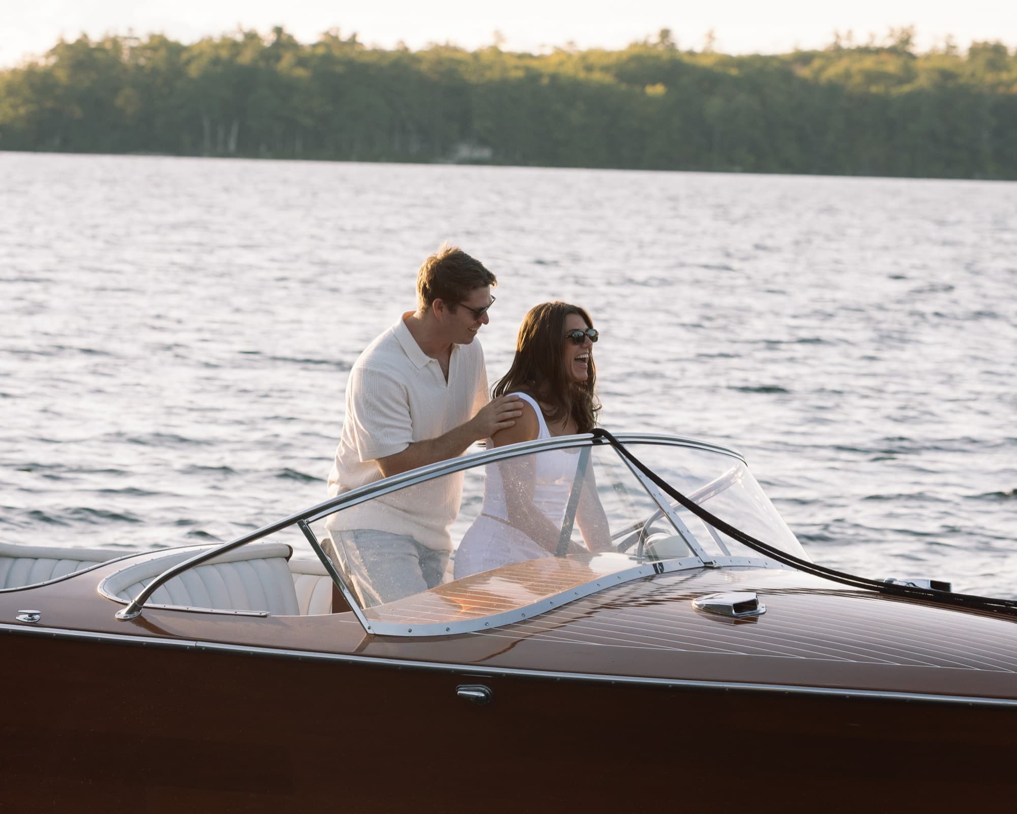 Morgan laughing beside Jon at the wheel of the runabout, both in sunglasses, the far shoreline soft behind them.